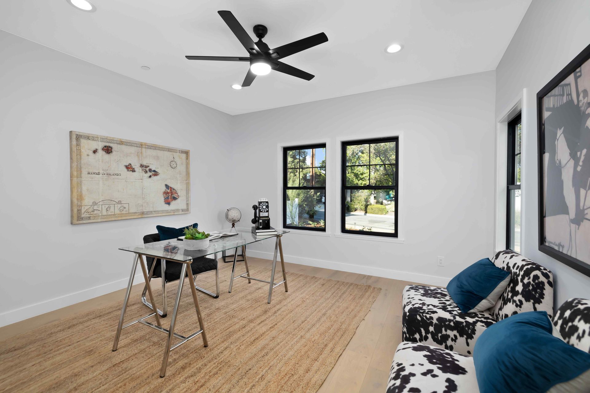 Home office with desk, rug, black-framed windows, cow print sofa, and a ceiling fan.