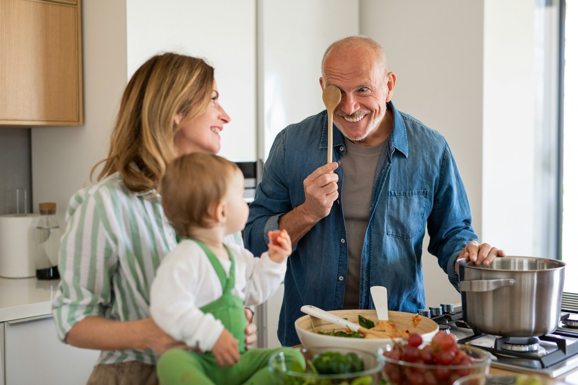 A woman is holding a baby and a man is cooking in the kitchen.