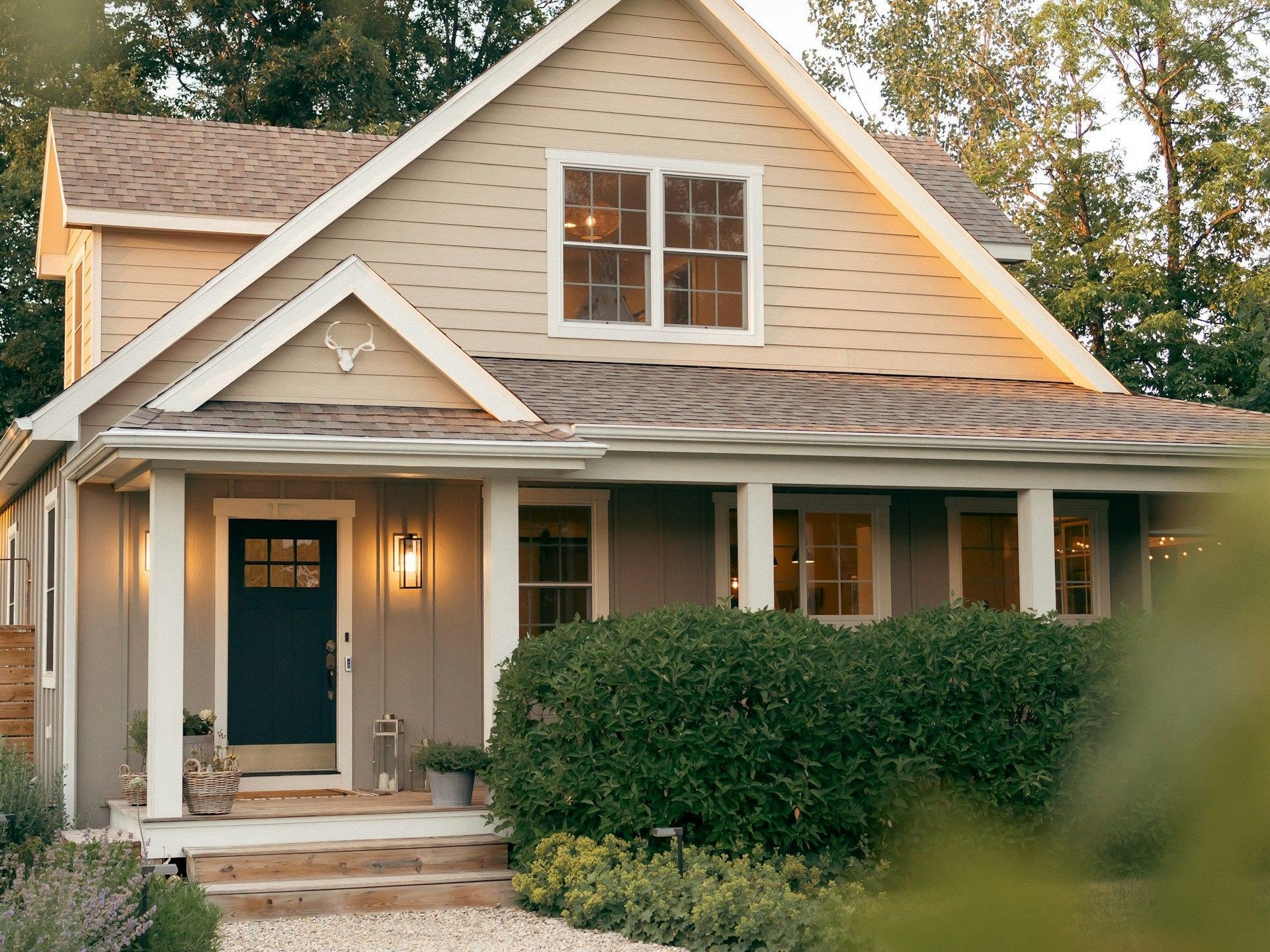 A house with a porch and a black door is surrounded by bushes and trees.