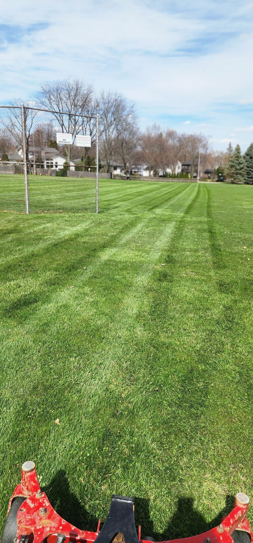 A green lawn being mowed with a riding lawnmower, showing visible stripes. Basketball hoops in the background.