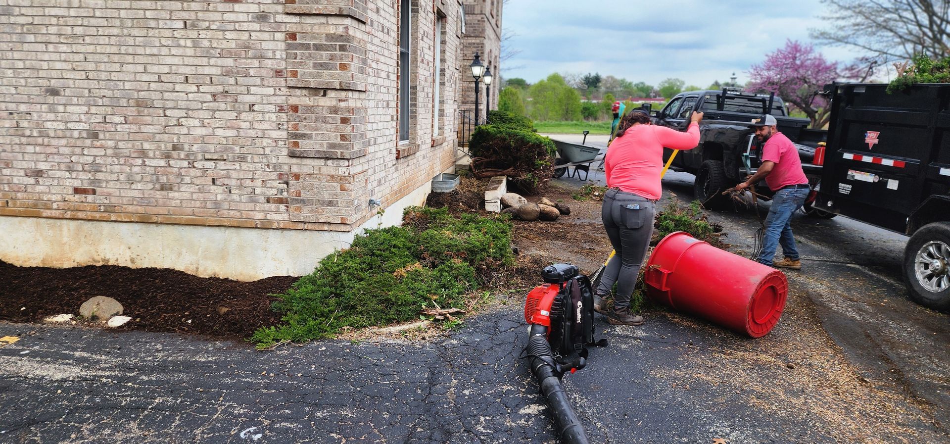Two people working outdoors by a brick building, using a leaf blower and trailer.