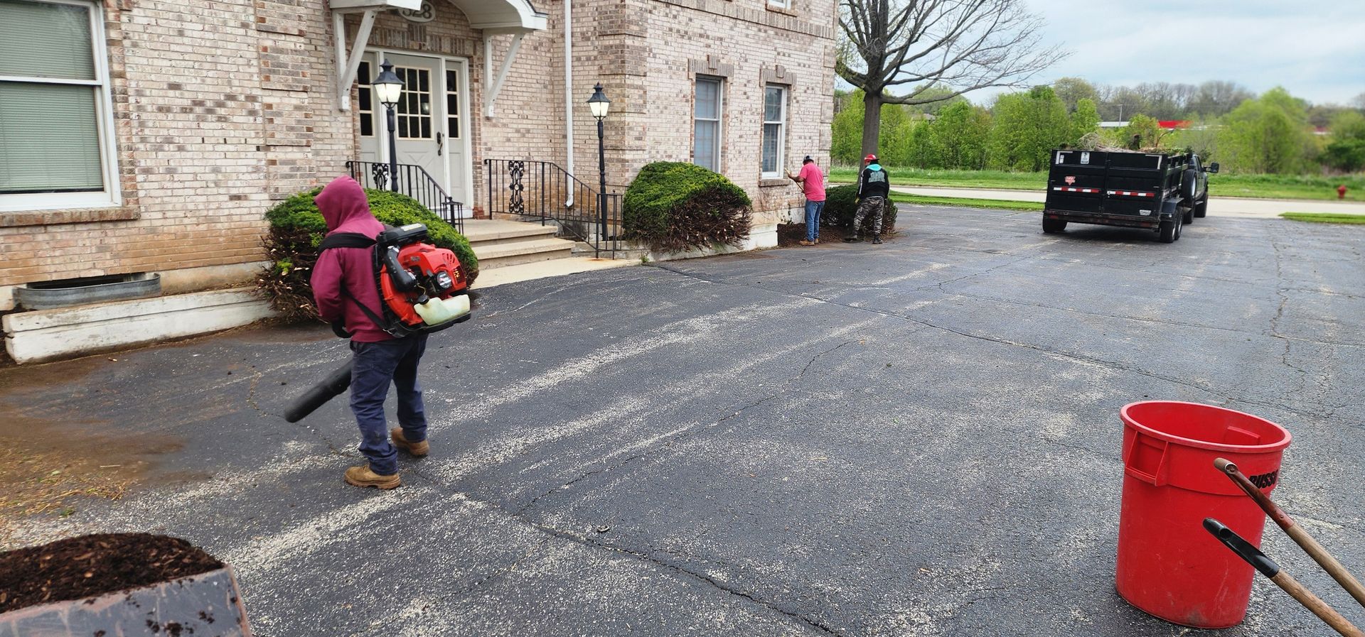 Person using leaf blower on a driveway, with a red bucket, truck, and building in the background.