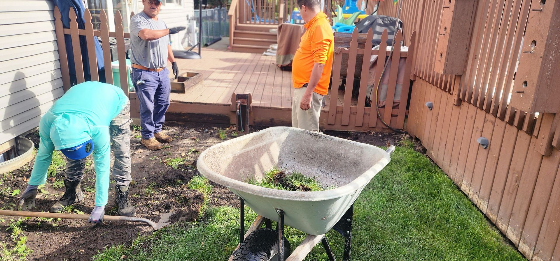 Three people working in a yard near a deck; one rakes soil, another observes, the third stands near a wheelbarrow.