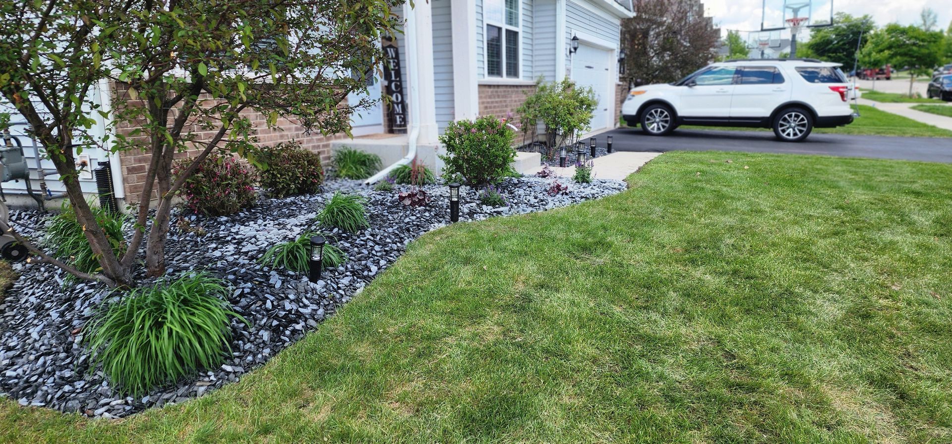 A house with a white SUV parked in the driveway. Landscaping includes green grass and rock beds.