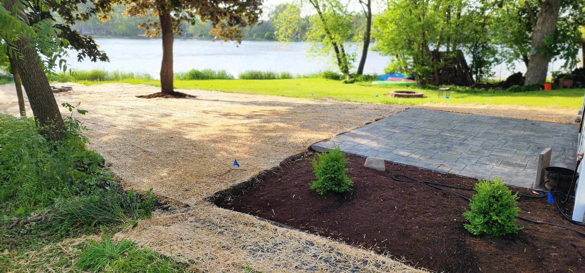 A lakeside yard with gravel, mulch, small bushes, and a concrete patio.
