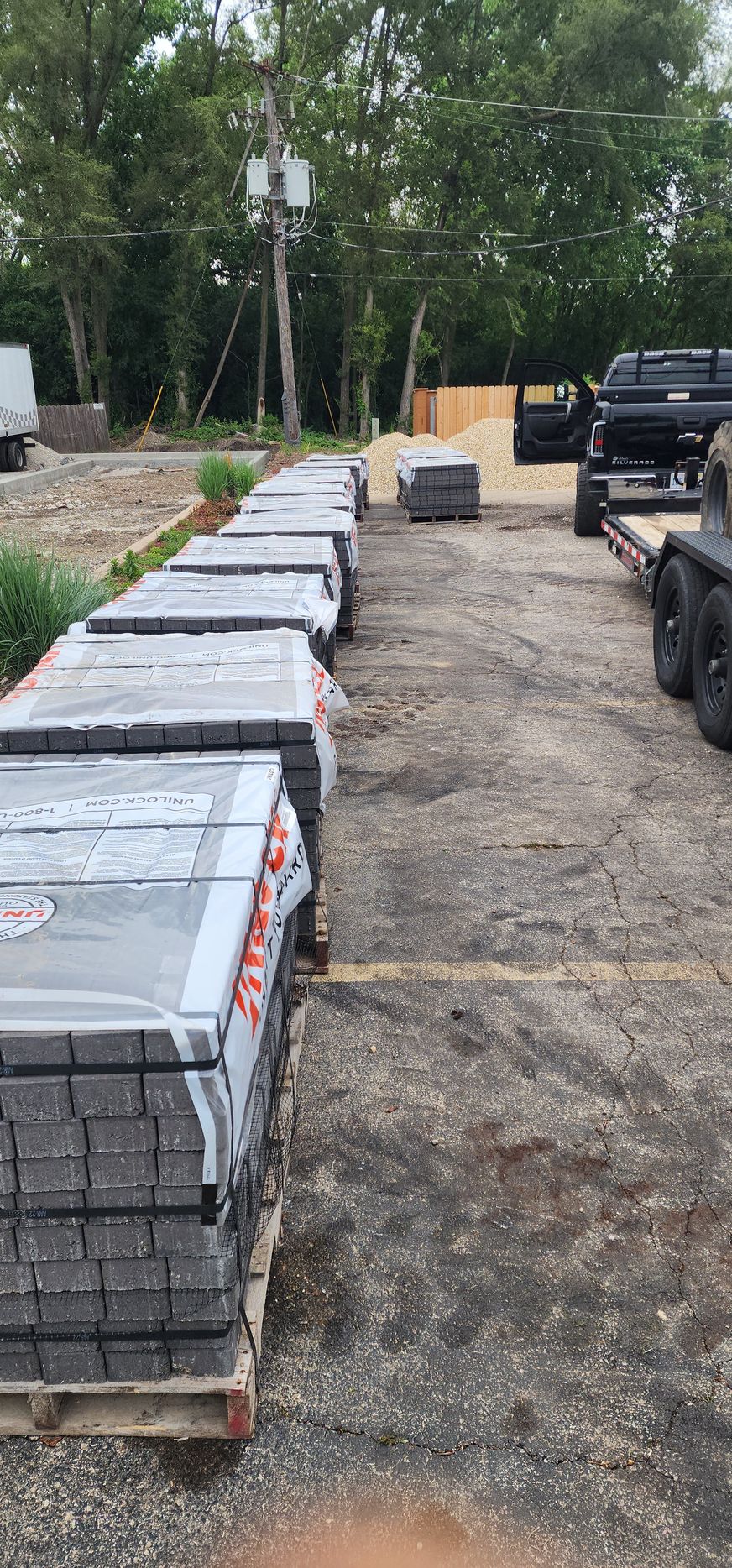 Pallets of paving stones stacked on a gravel lot near trees and a trailer.