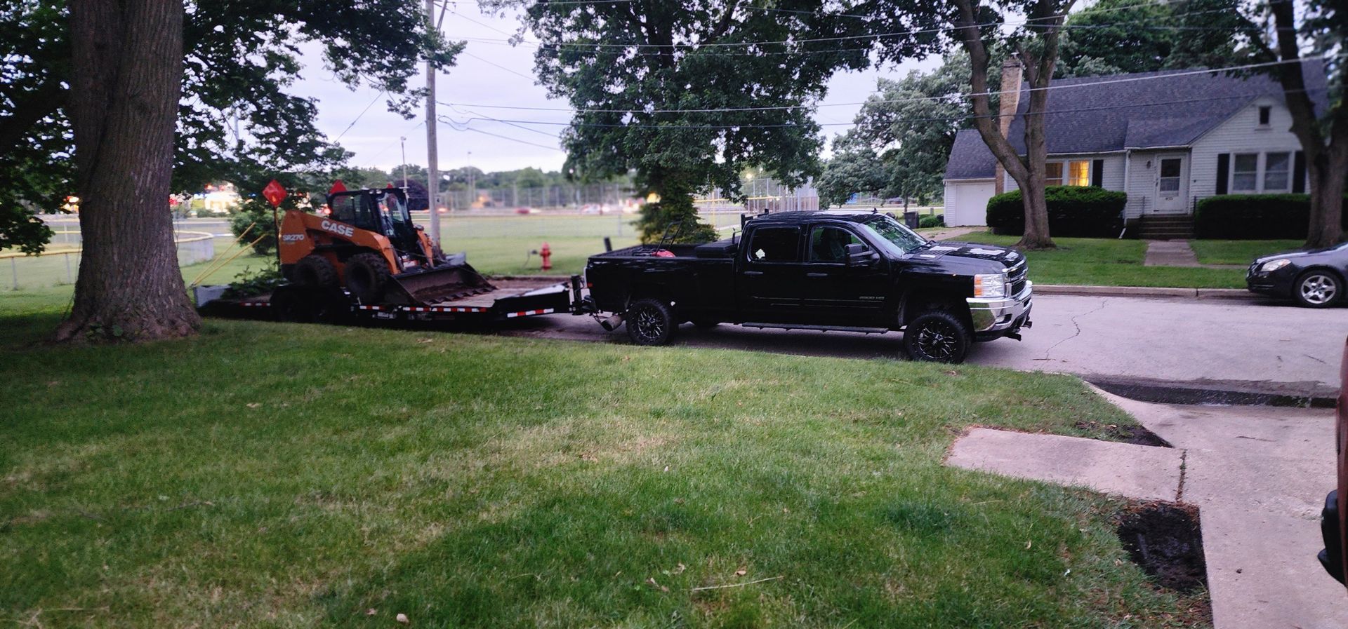 Black pickup truck towing an excavator on a trailer in a residential area with green lawn and a house.