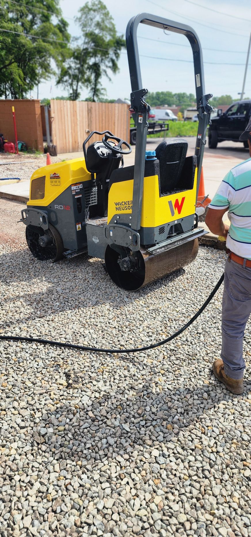 A yellow and gray road roller on a gravel surface with a person holding a hose.