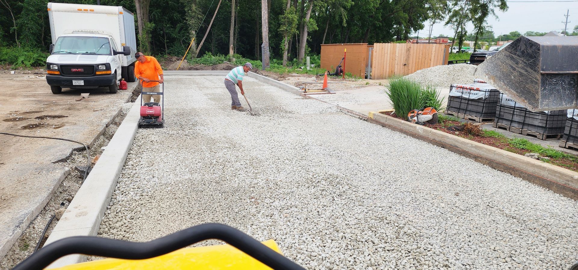 Construction workers paving a gravel driveway. One uses a compactor, another rakes gravel. A truck and a wooden fence are visible.