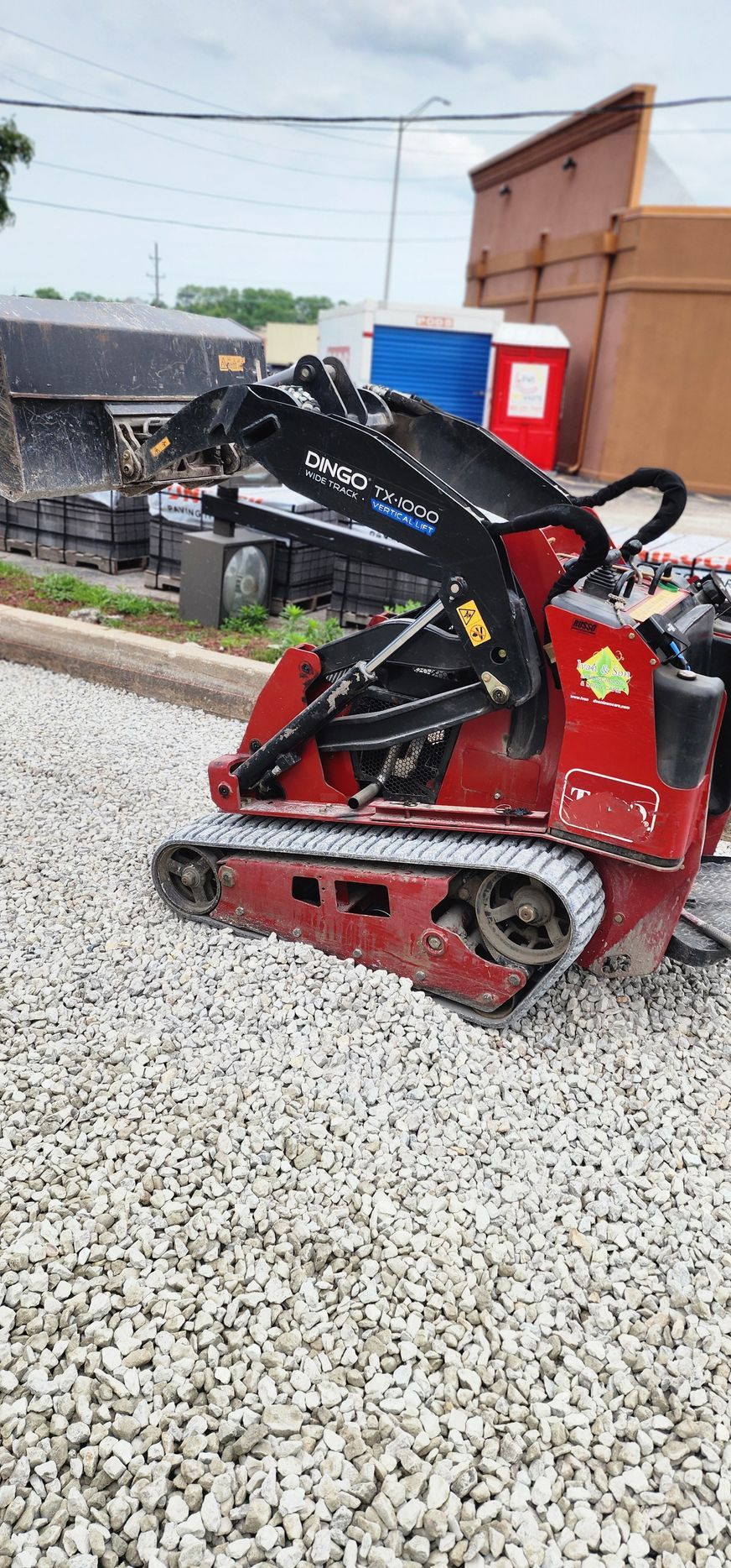 Red tracked mini skid steer on a gravel surface next to a sidewalk and construction materials.