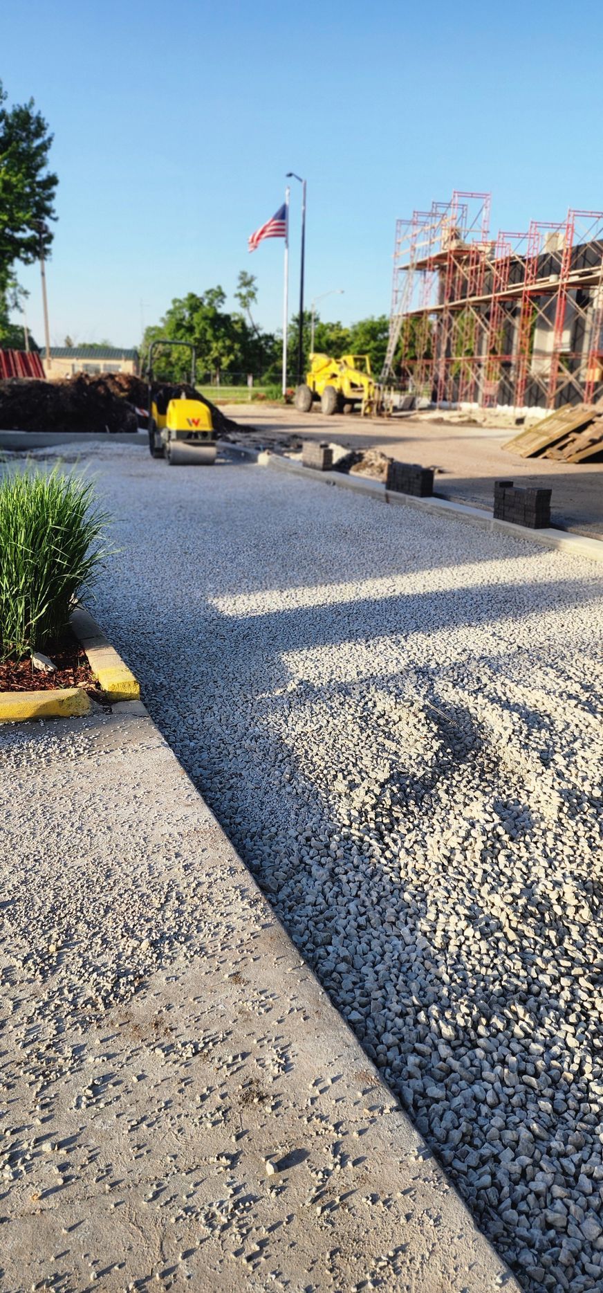 Construction site with gravel pathway, small compactor, American flag, and building.