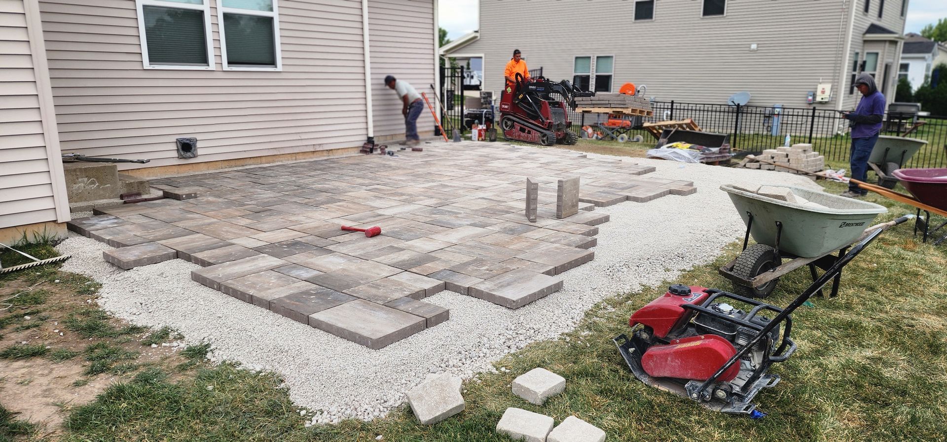 Workers building a stone patio in a backyard; a wheelbarrow, compactor, and gravel are visible.