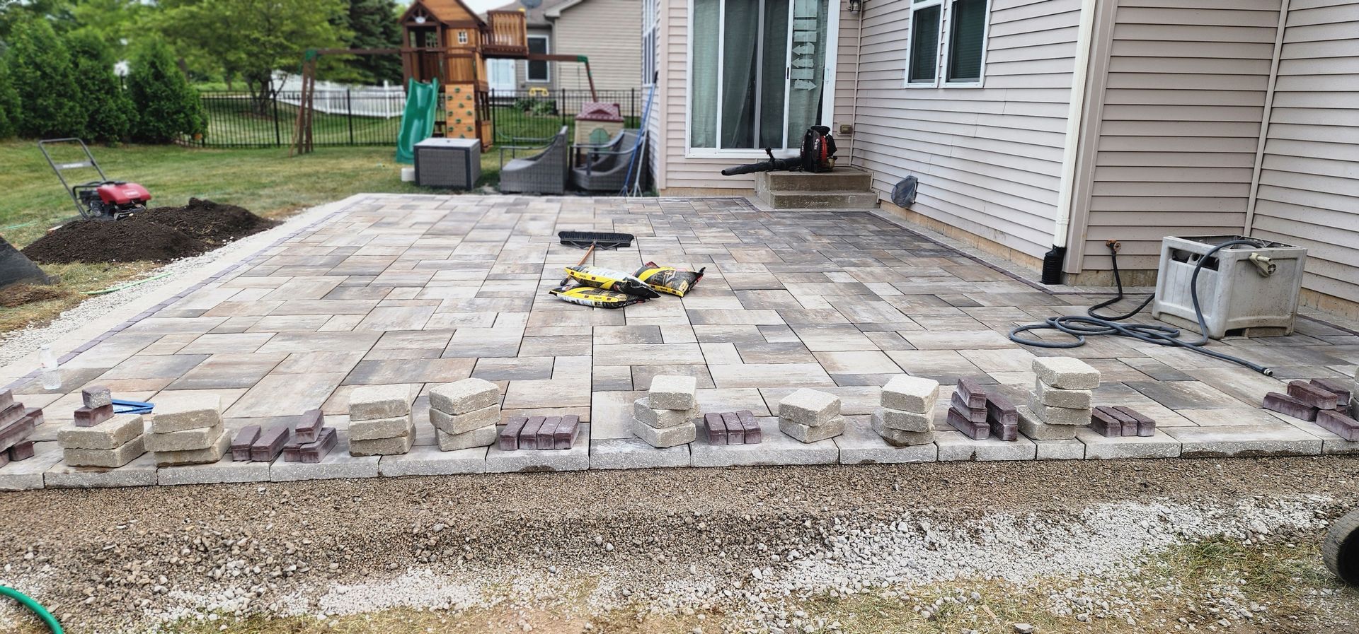 Patio construction in progress with pavers and tools. Gravel base and house in the background.