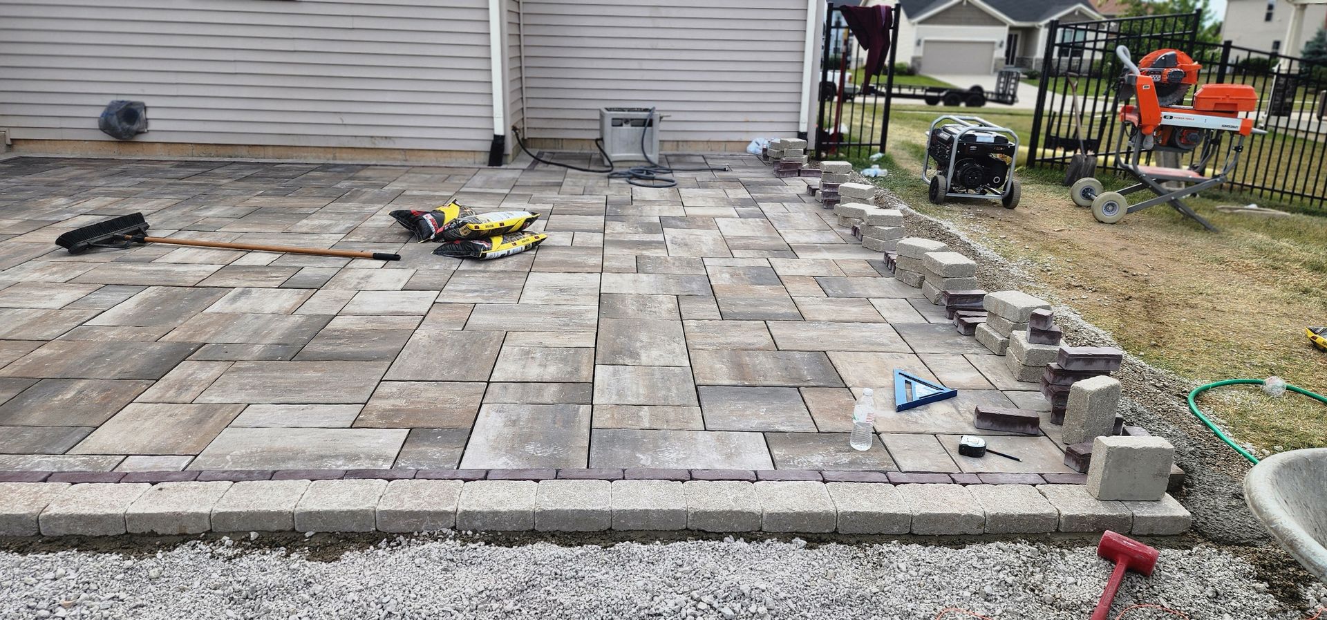Patio installation in progress with pavers, edging, and tools in a yard.