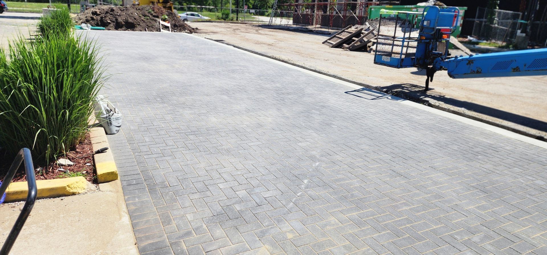 A paved area with plants and a construction lift. Construction materials in the background. Sunny day.