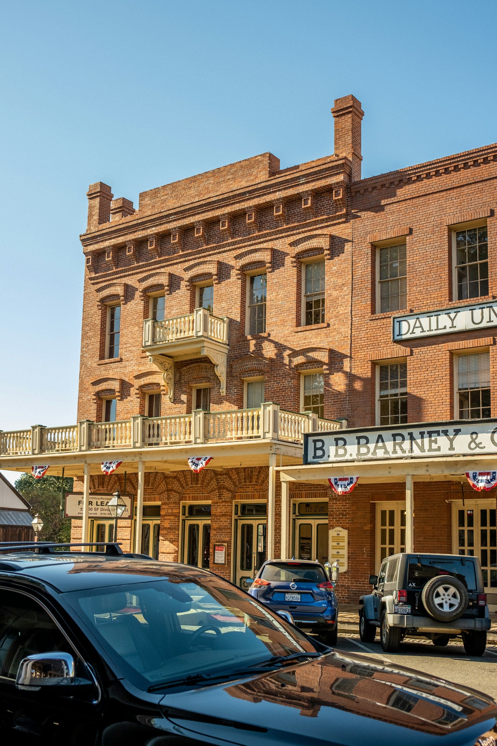 Historic brick building with balconies and awnings. Cars parked in front, clear blue sky.