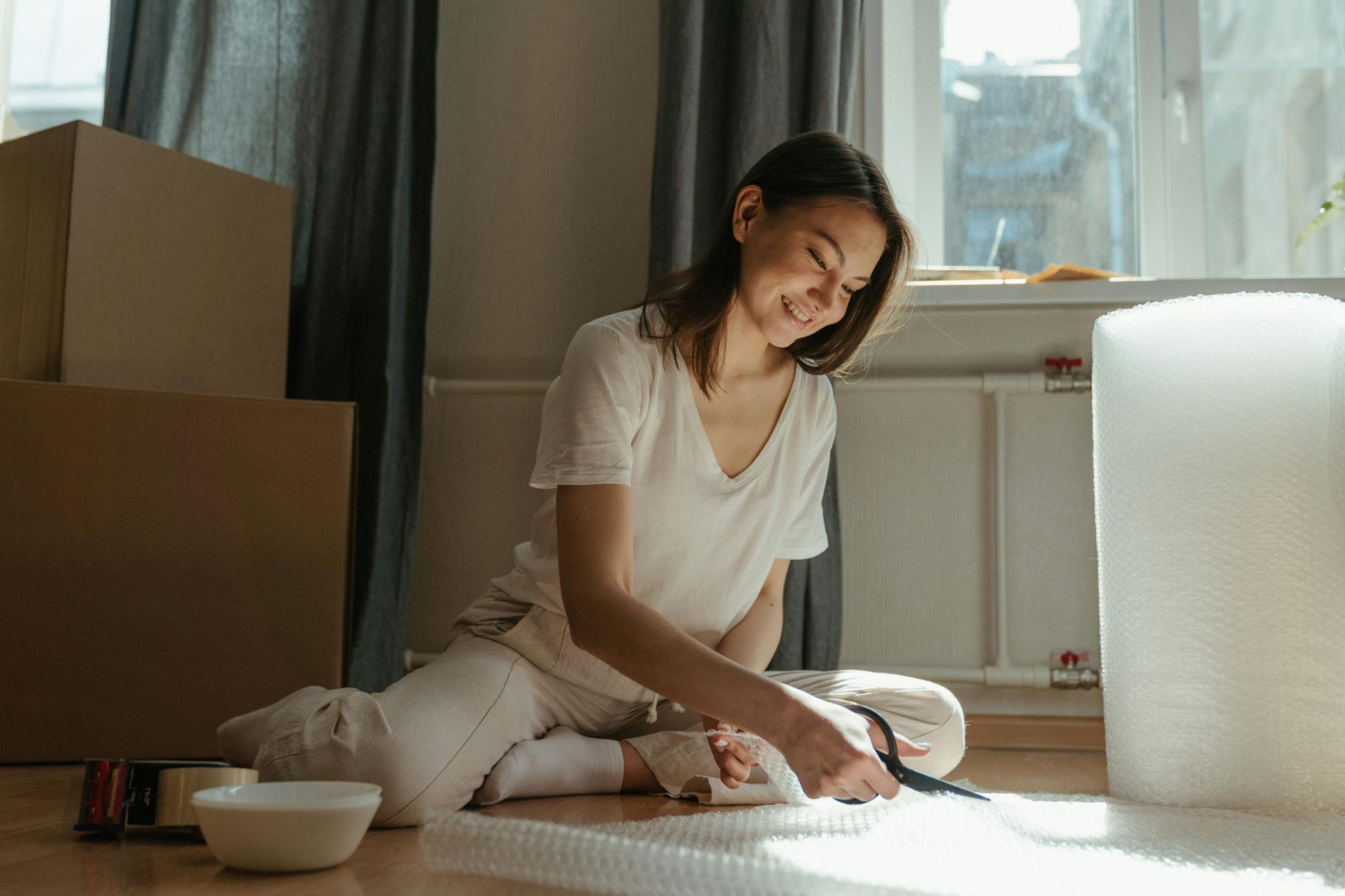 Woman in casual clothes sits on the floor cutting bubble wrap with scissors, smiling, boxes nearby.