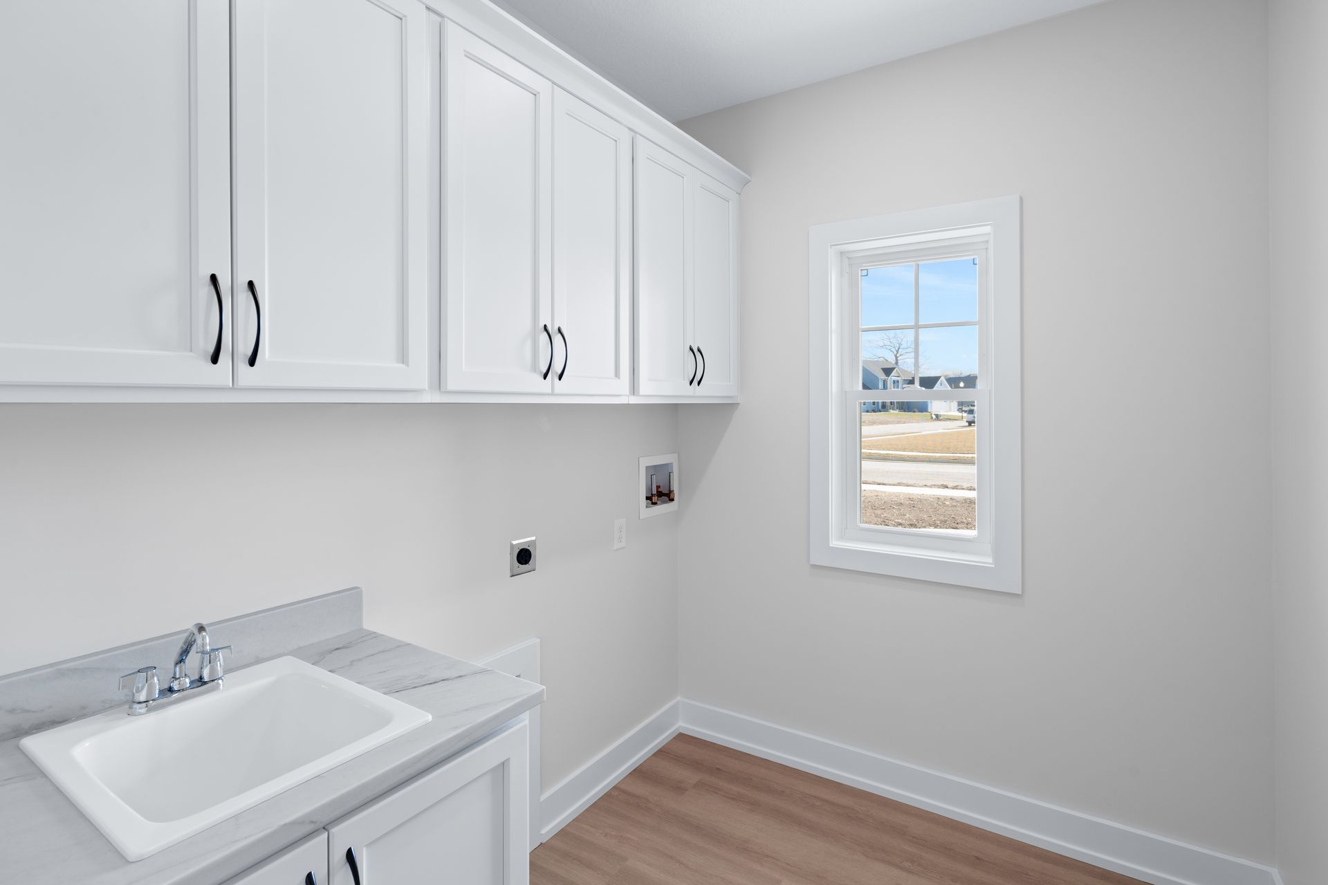 A laundry room with white cabinets , a sink , and a window.