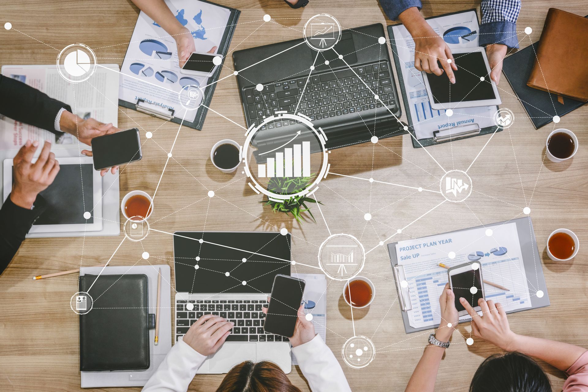 A top-down view of people working at a wooden table with laptops and documents, overlaid with digital data icons.