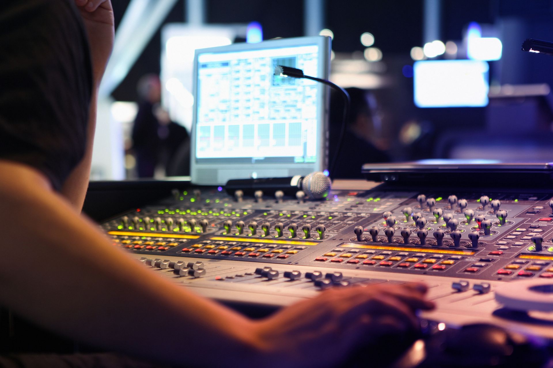 person operating a professional audio mixing console and computer screen displaying audio software is visible in the background