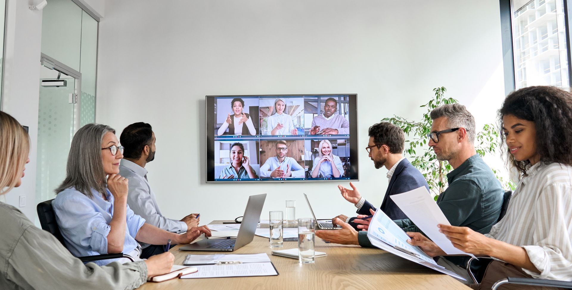 Business meeting in a conference room with six people on a video call displayed on a wall monitor