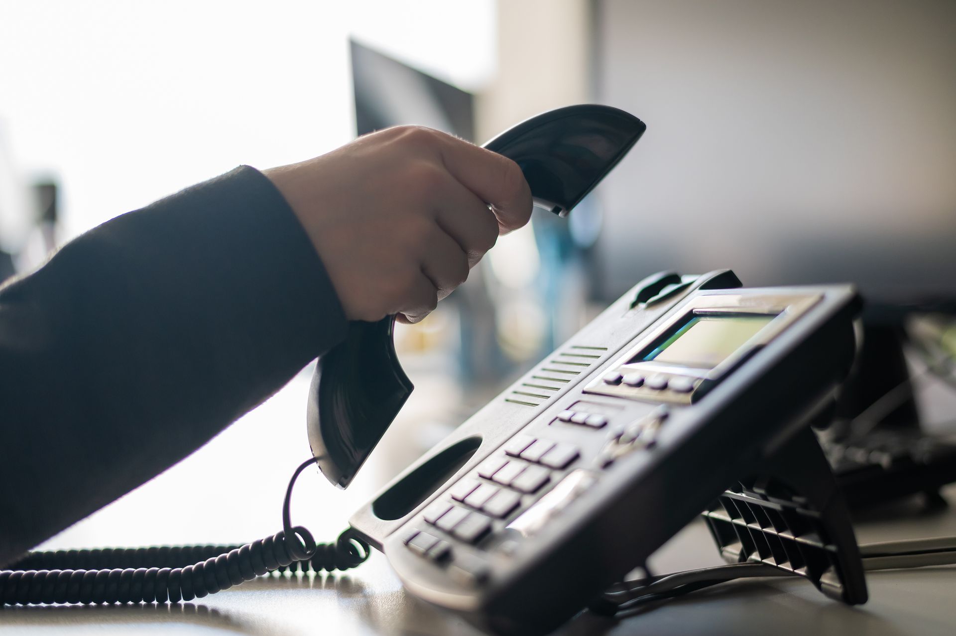 A person holding the receiver of a black office desk phone in a professional, brightly lit workspace.