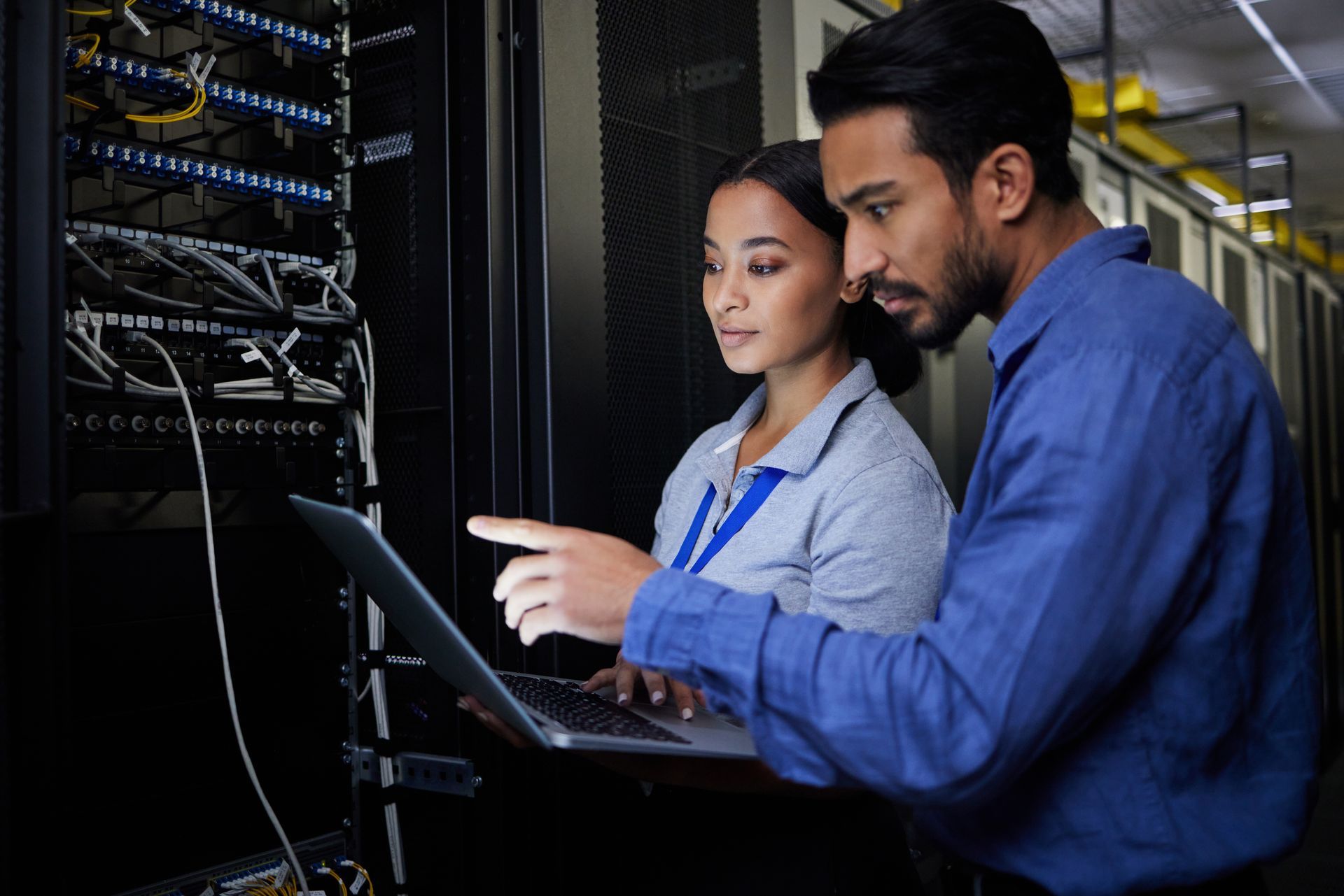 Two IT professionals collaborate on a laptop while inspecting the wiring and hardware inside a server room.