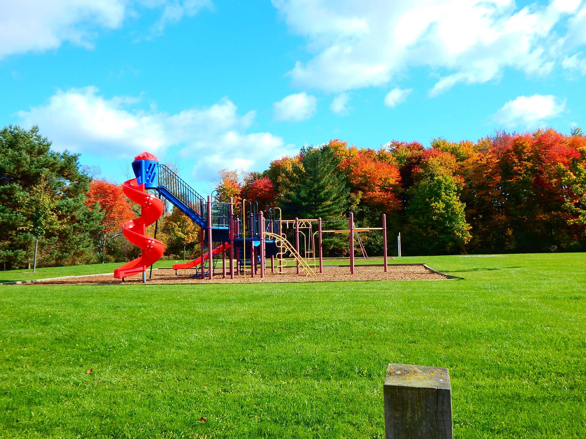 A playground with a red slide in the middle of a grassy field.