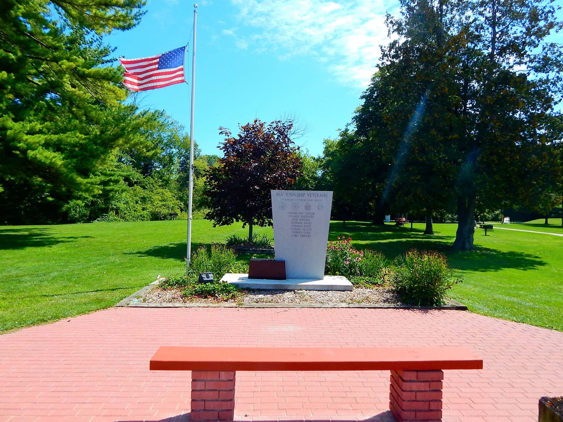 A brick bench sits in front of a memorial with an American flag in the background.