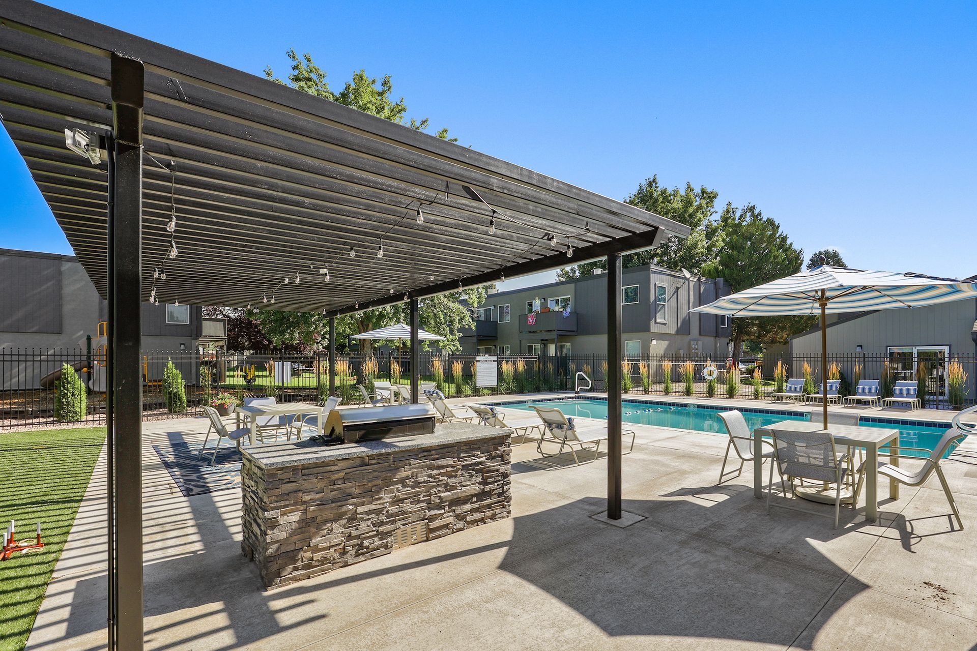 Outdoor pool area with a stone grill island under a pergola, lounge chairs, and a table with an umbrella on a patio at Alpine Lofts in Windsor, CO.