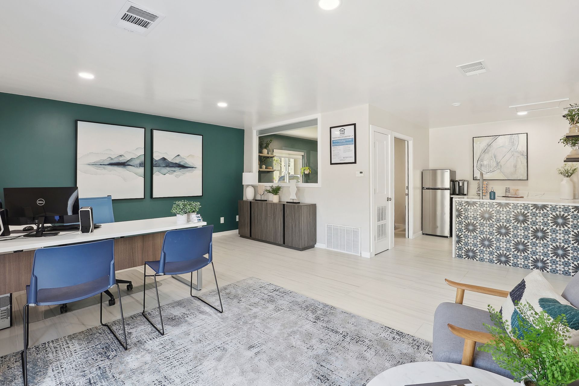 A modern office space with a teal accent wall, white desk, blue chairs, a patterned kitchen island, and a gray rug at Alpine Lofts in Windsor, CO.