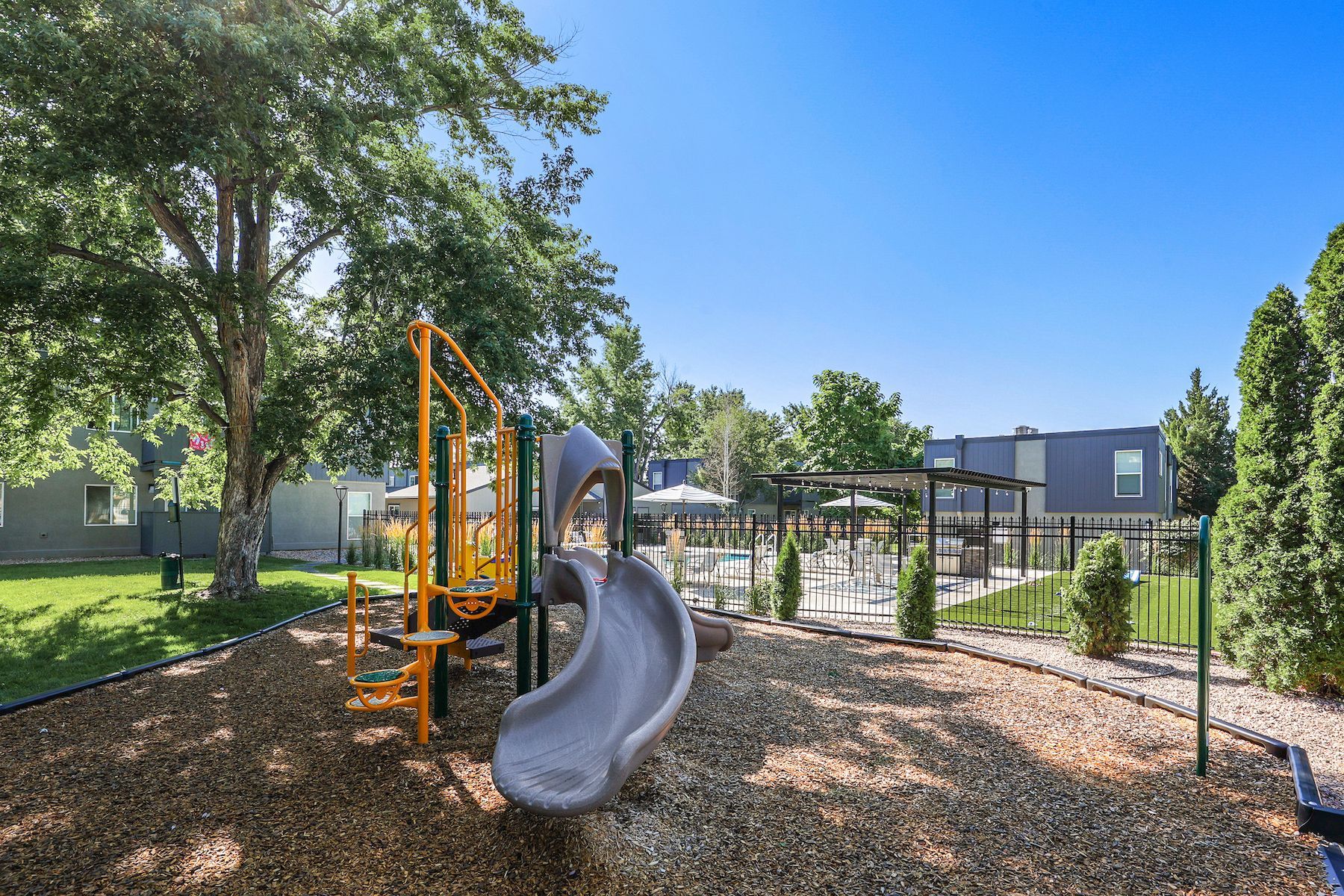 A playground with a grey slide and yellow climbing frame on wood chips, with a pool and apartment buildings in the back at Alpine Lofts in Windsor, CO.