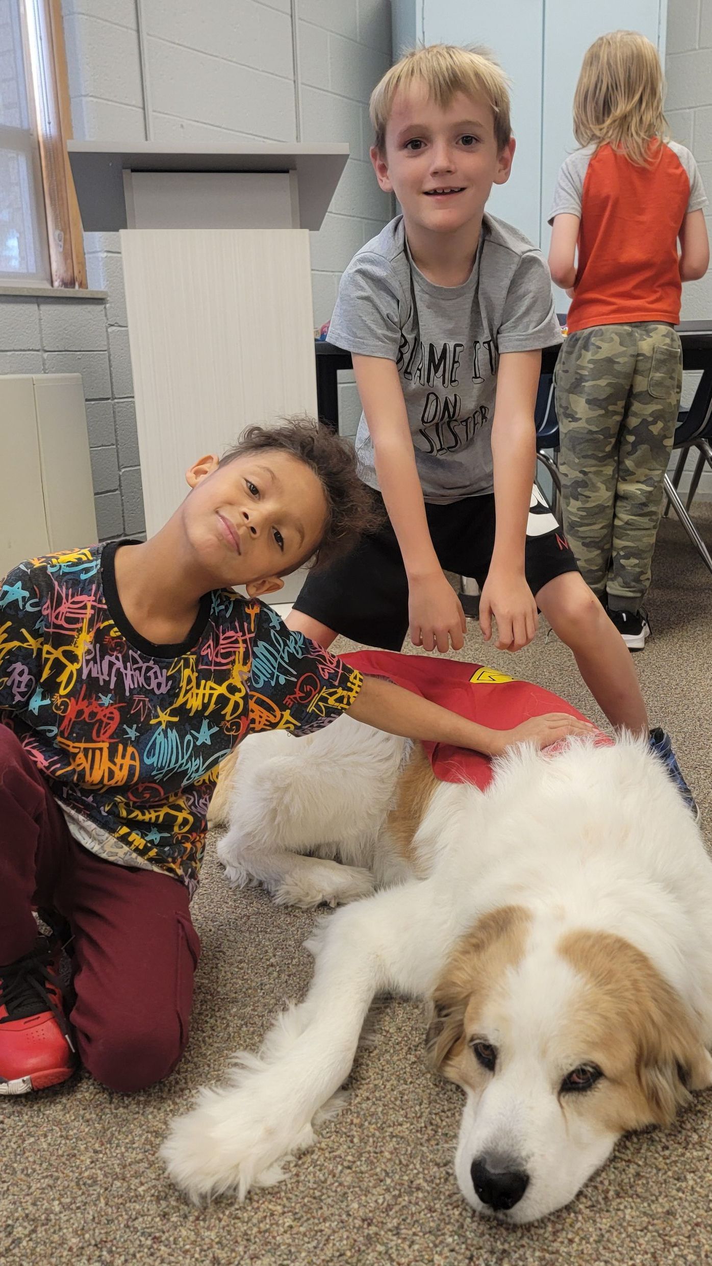 Two young boys are kneeling next to a dog in a classroom.