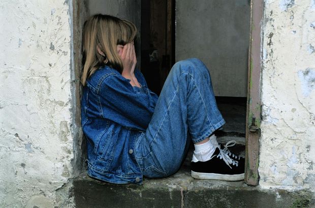 A girl in a denim jacket is sitting in a doorway covering her face with her hands.