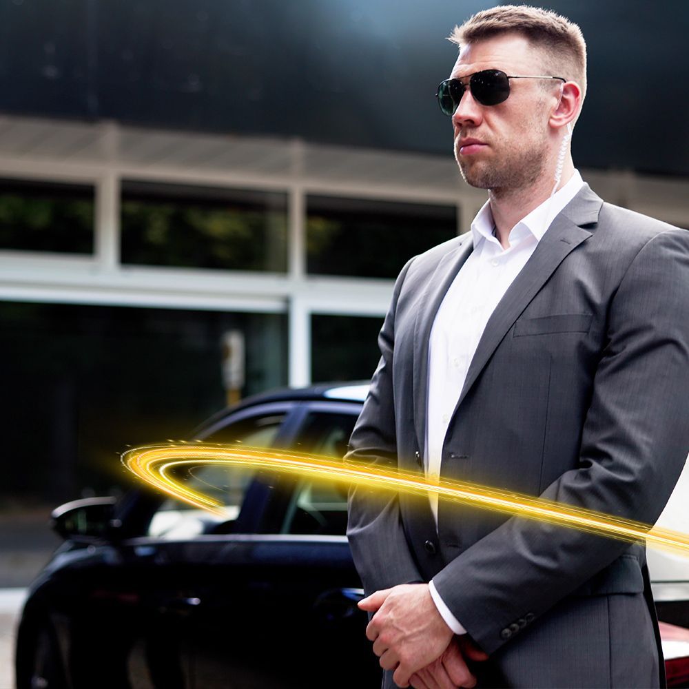 Man in suit and sunglasses, standing guard near a black car, with a glowing yellow trail.