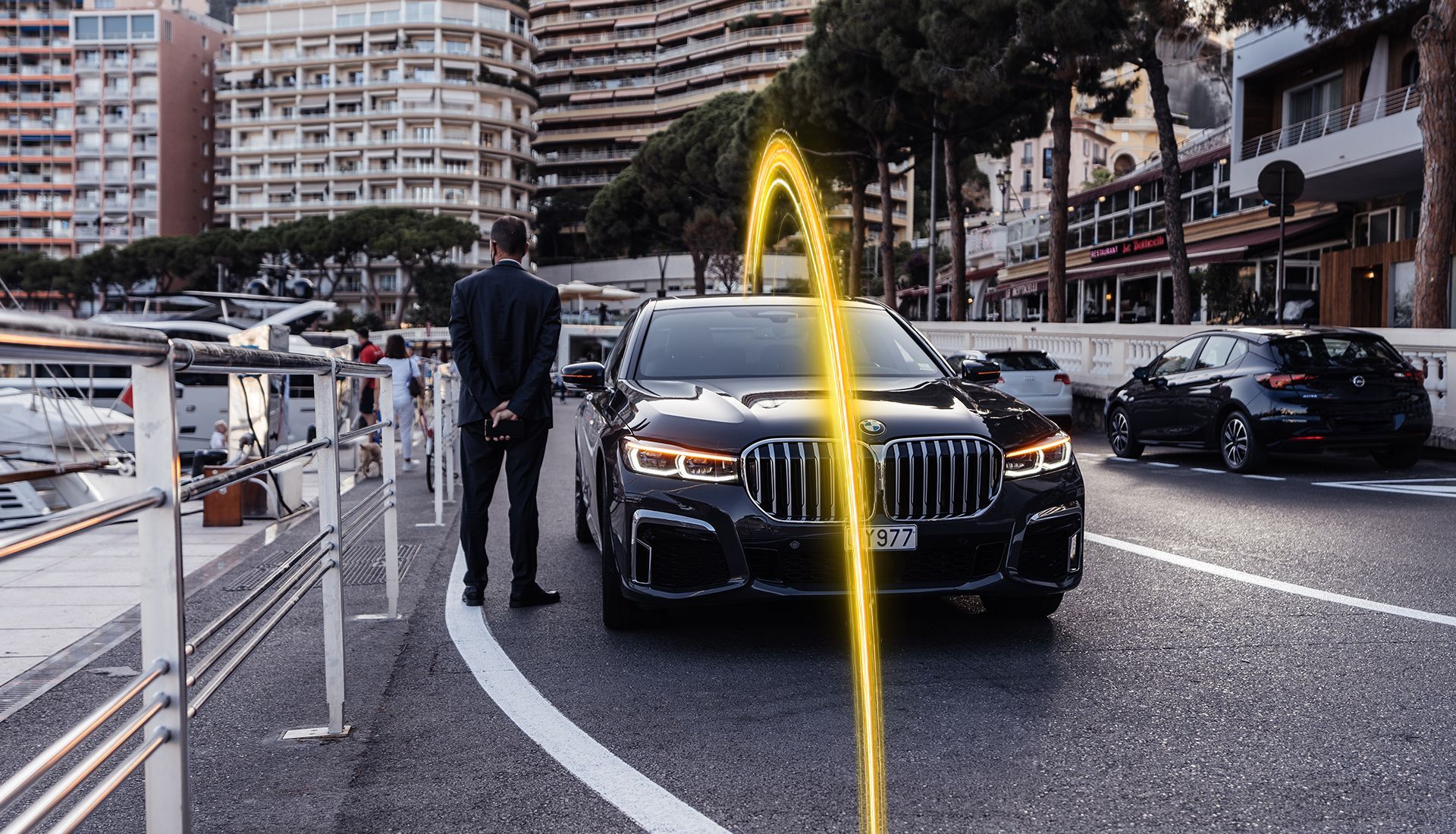 Man in black suit stands by a black car with a yellow energy arc in Monaco.