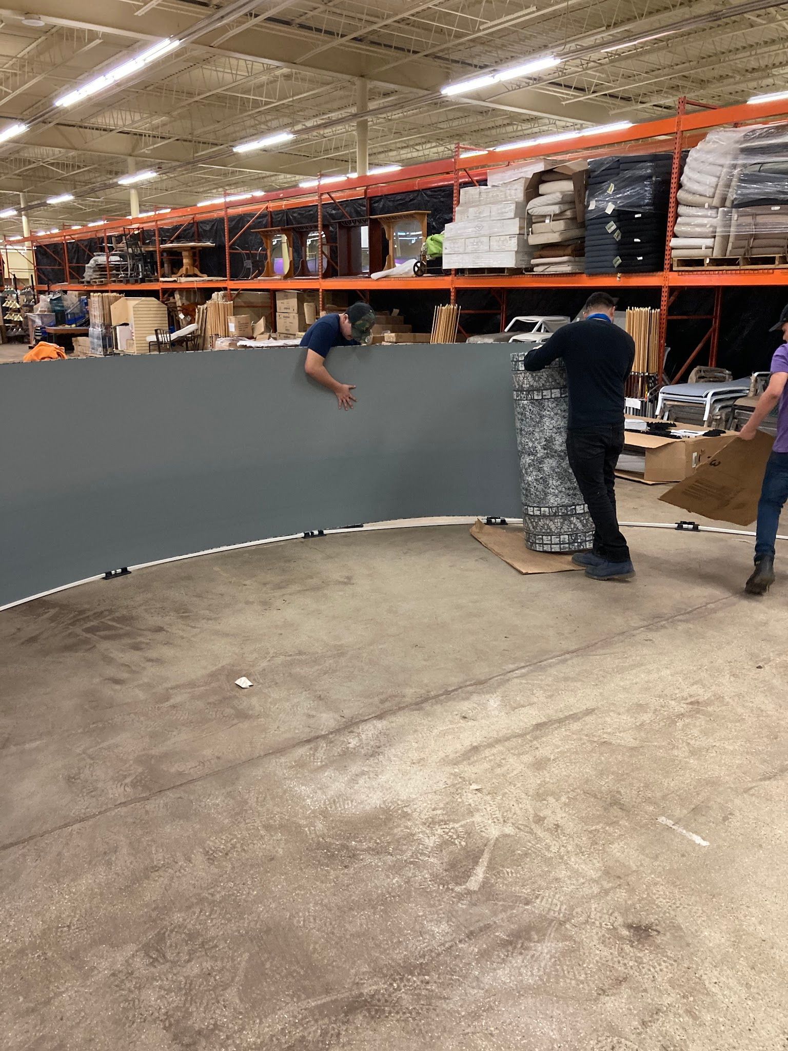 Workers moving a large gray curved panel in a warehouse. One is climbing on it, others are assisting.