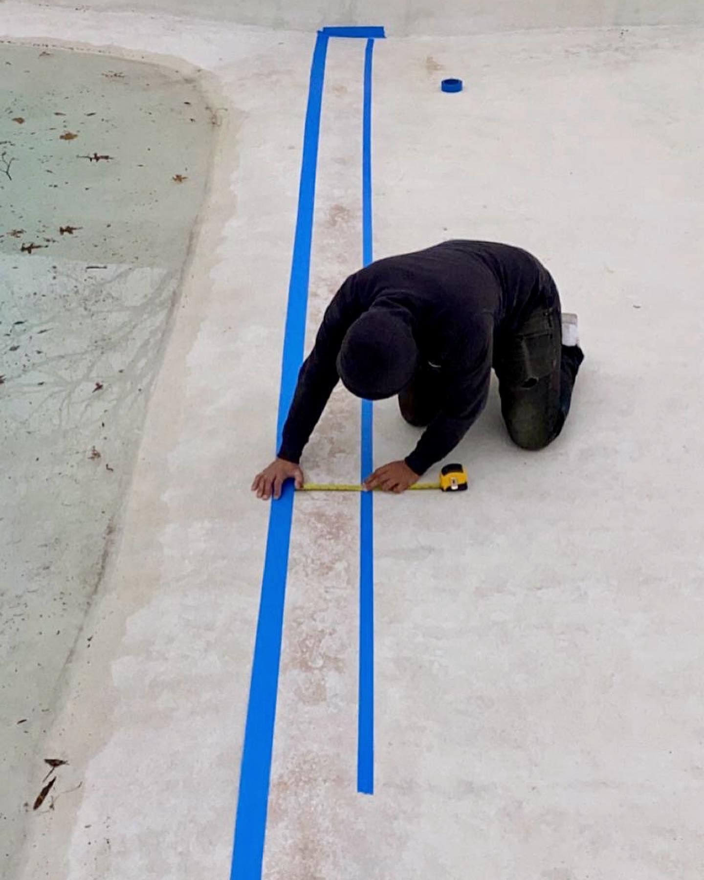 Person kneeling by a pool, measuring between two blue taped lines with a tape measure.