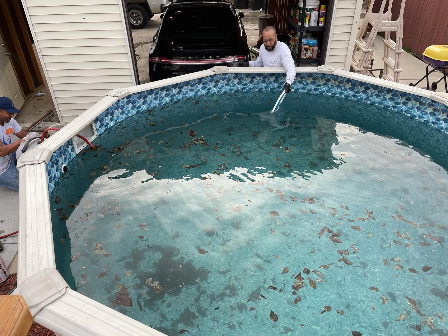 Two people cleaning a dirty above-ground pool with debris. One uses a net, other works on a hose.