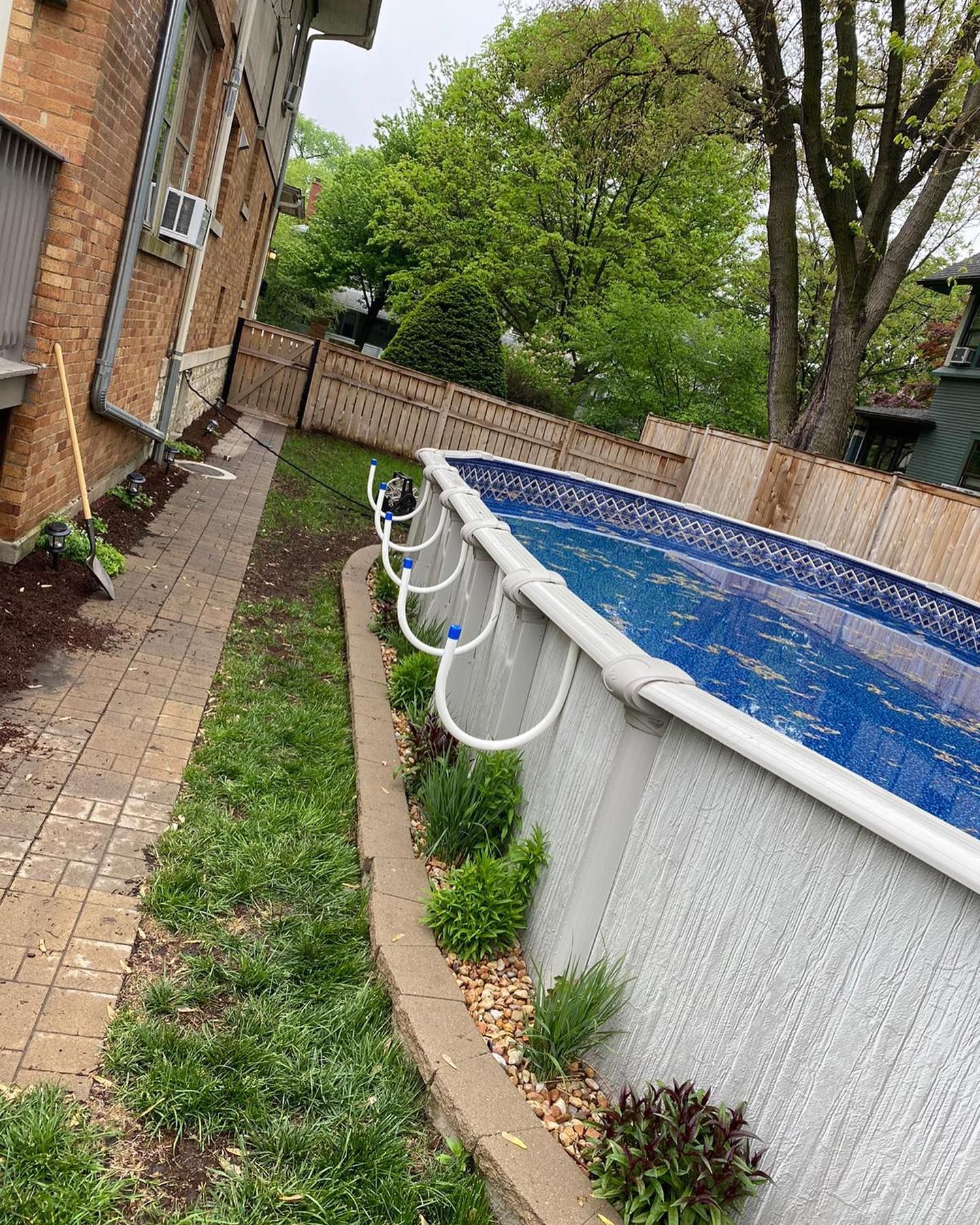 Above-ground pool next to a brick building and a narrow path with plants. Fence and trees in the background.