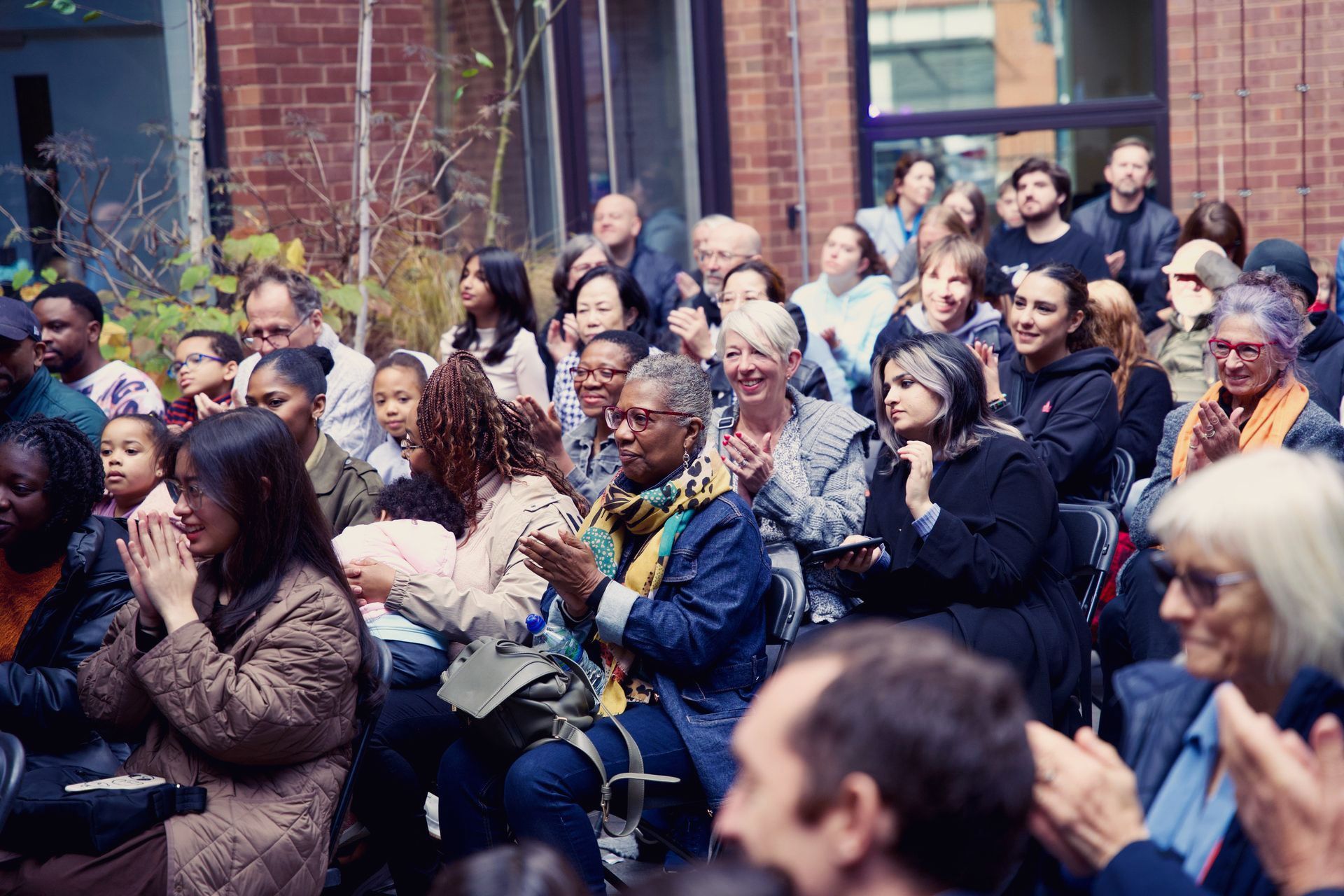 A large group of people are sitting in front of a brick building applauding.