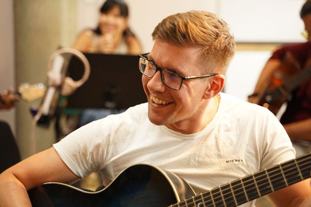 A man wearing glasses is playing a guitar and smiling.