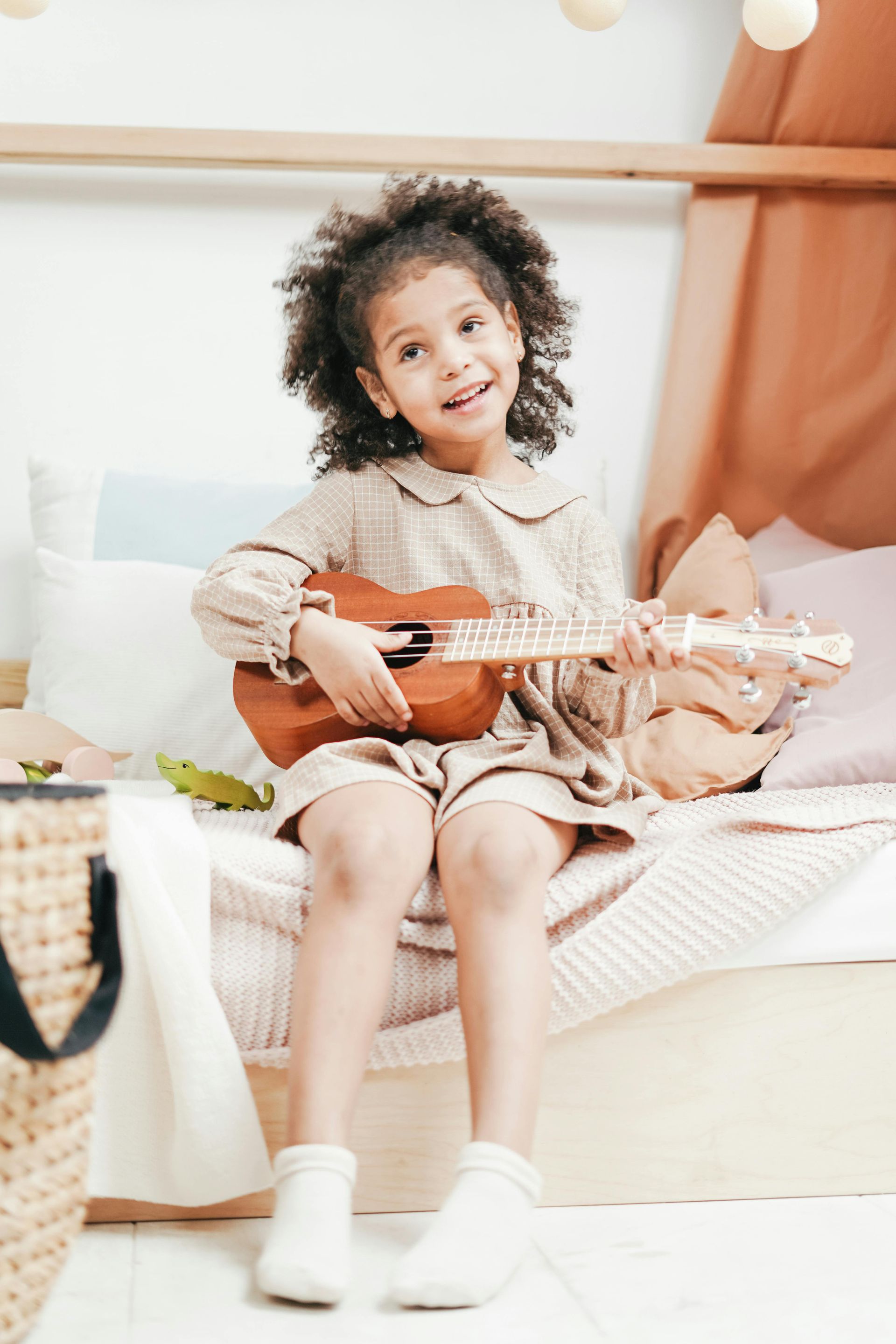 A little girl is sitting on a bed playing an ukulele.