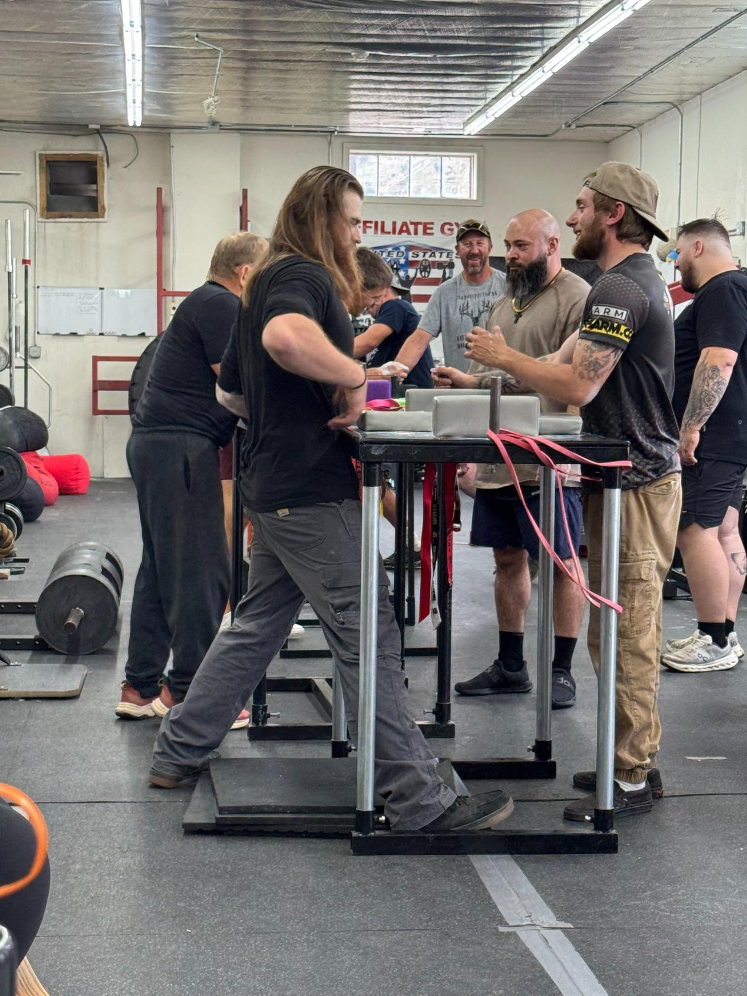 People arm wrestling at a table in a gym. Competitors are focused, with spectators watching.