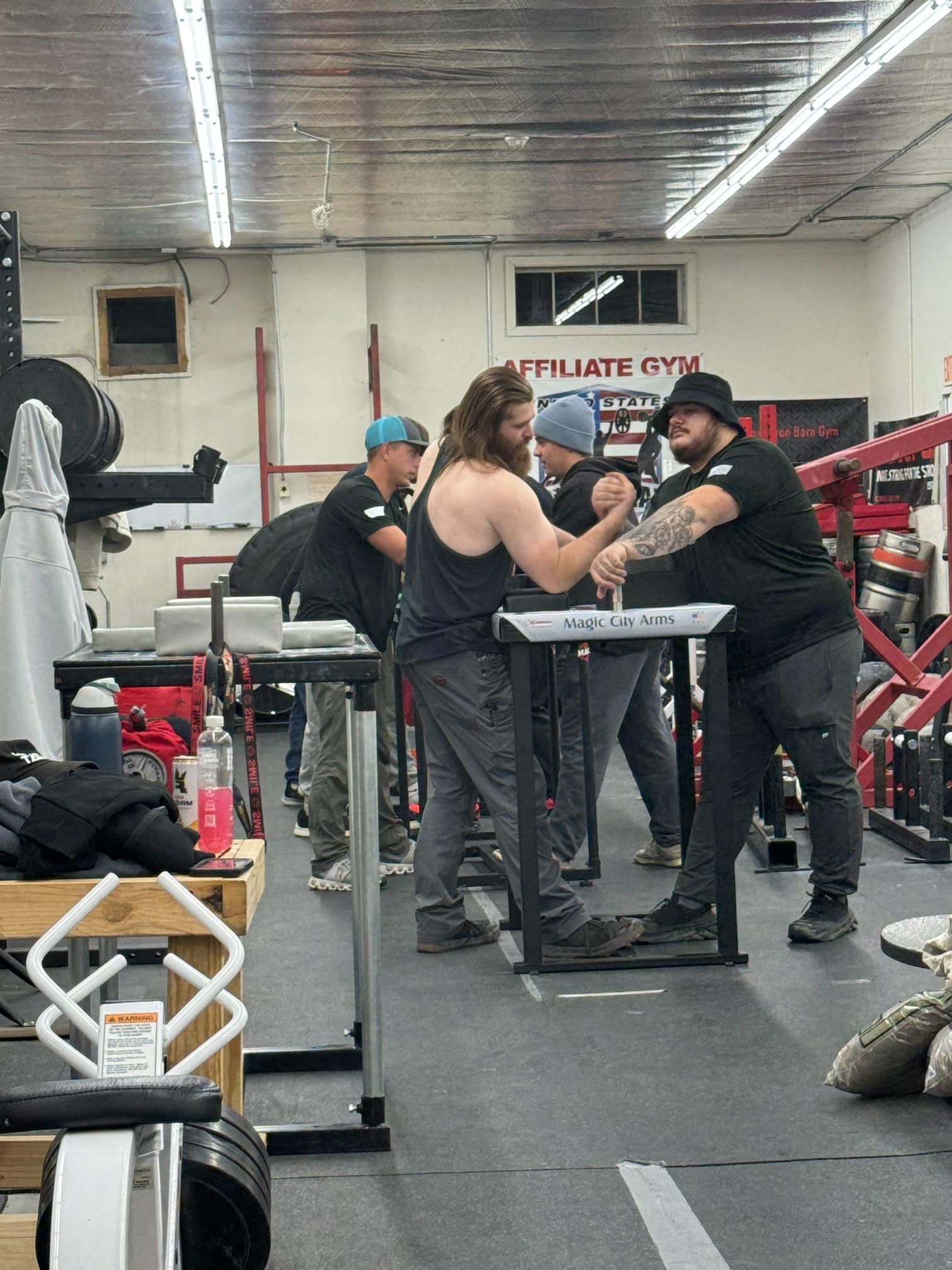 Two men arm wrestle at a gym; others watch. They stand at a table, both with hands clasped.