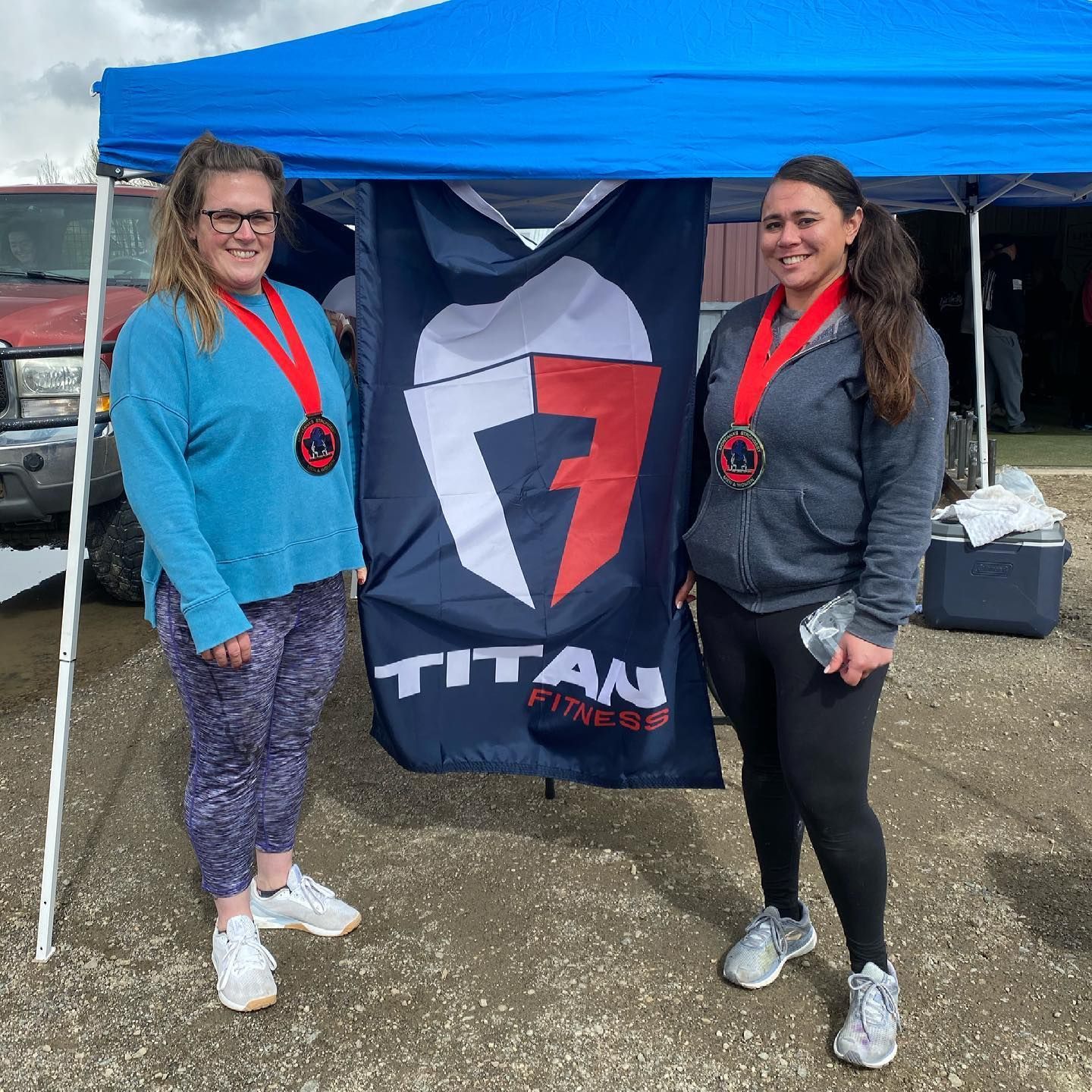 Two women in athletic wear with medals pose in front of a Titan Fitness banner.