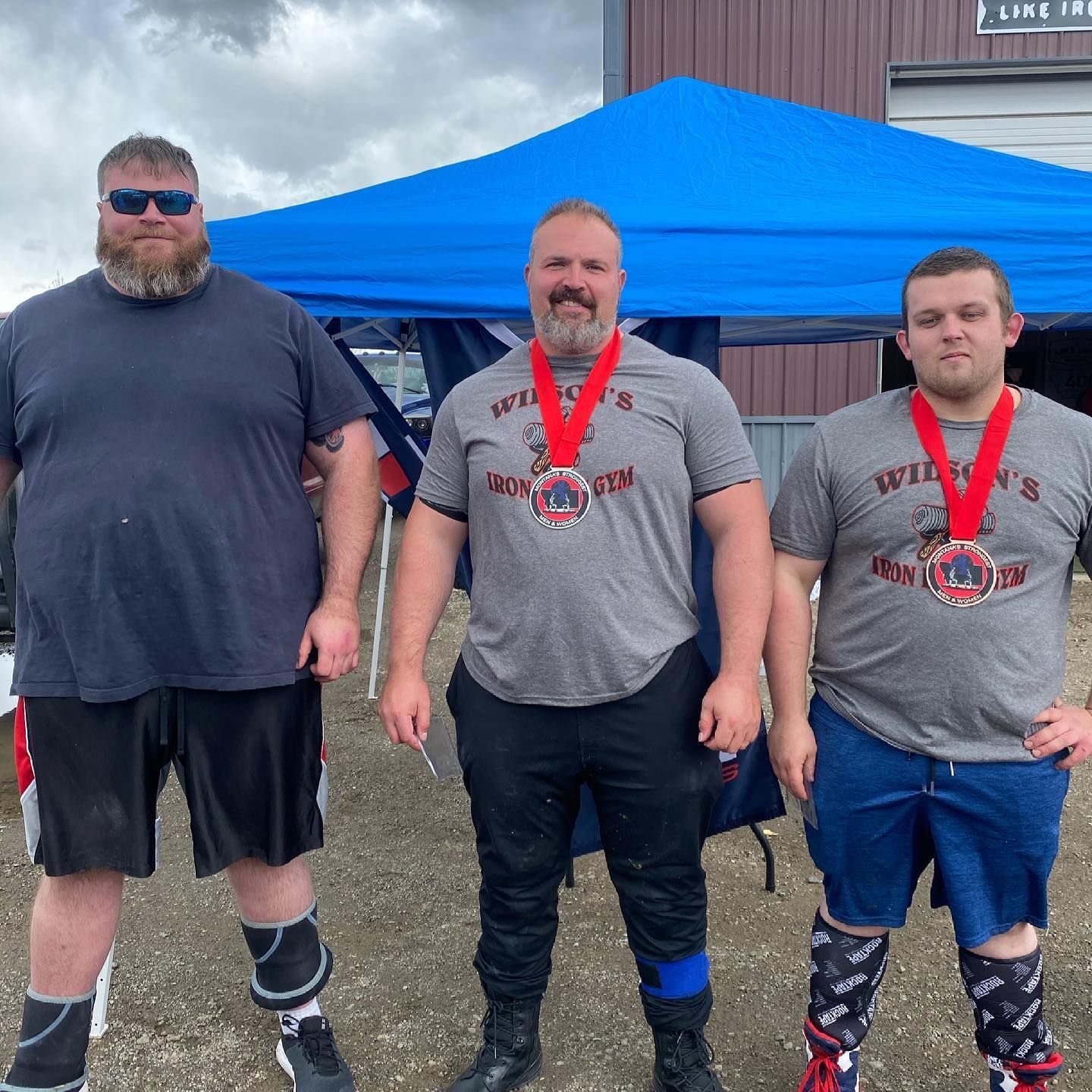 Three strongmen wearing medals stand under a blue canopy. They are posing after a weightlifting competition.