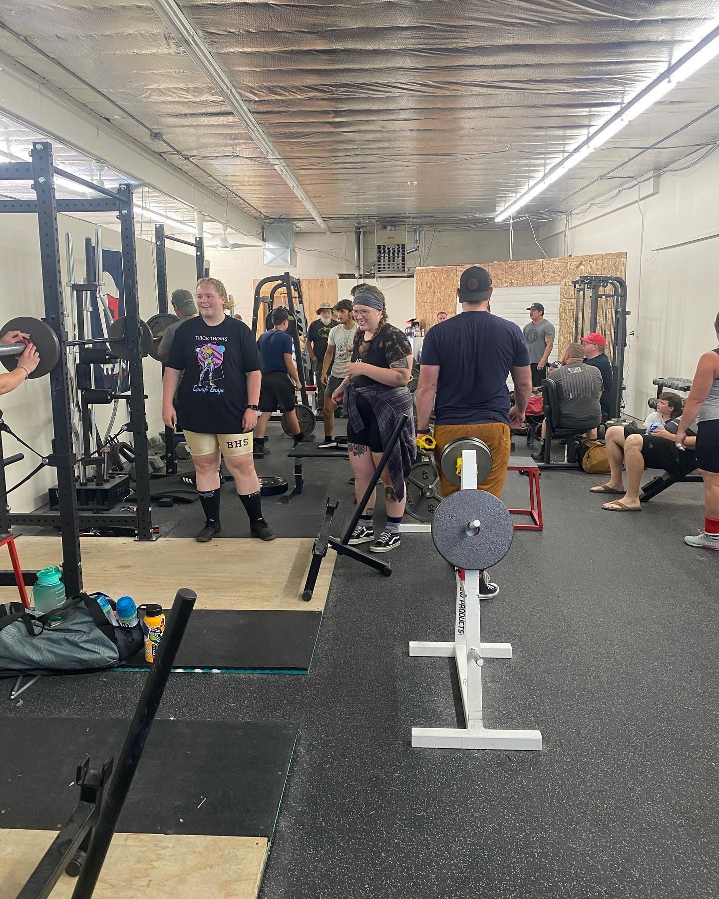 People at a weightlifting gym. Several are standing near equipment, some with weights. Dark gym setting.