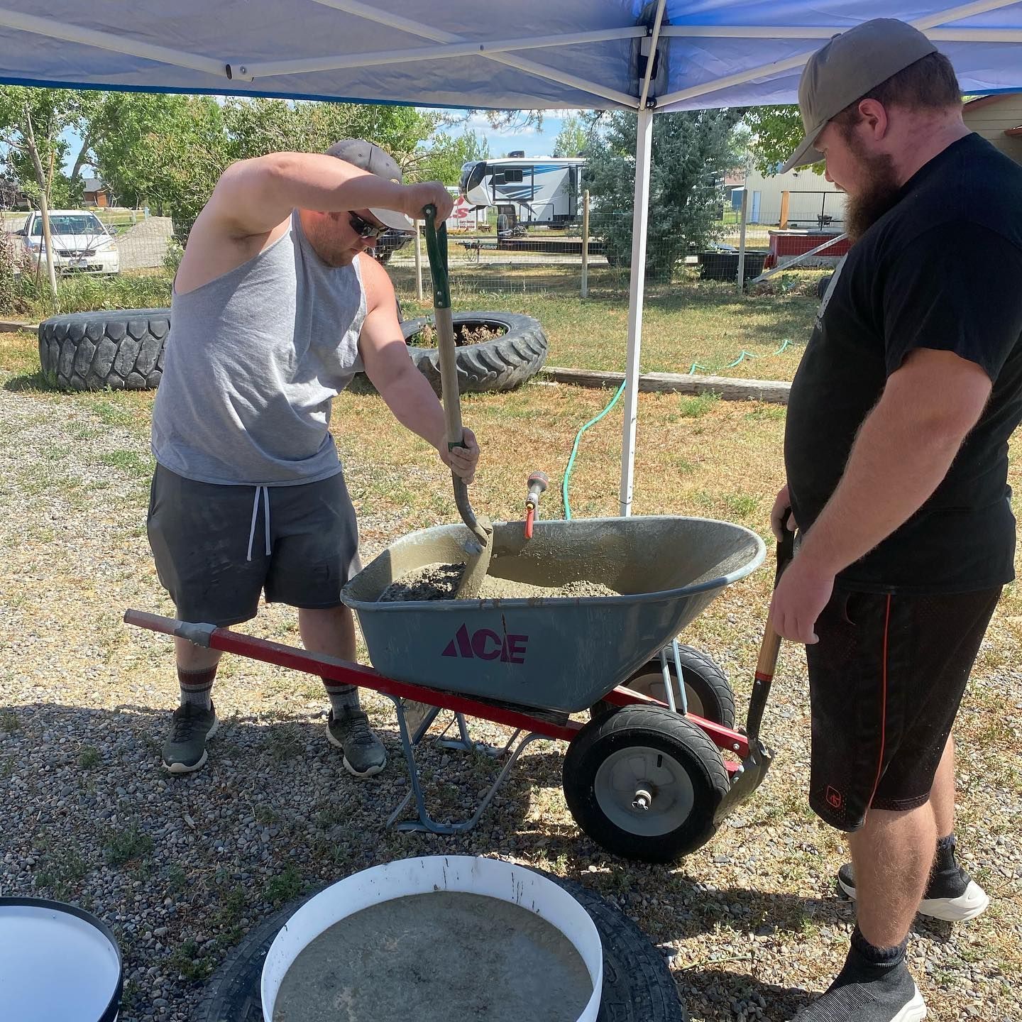 Two men mixing concrete in a wheelbarrow outdoors. One shovels, the other stands by.