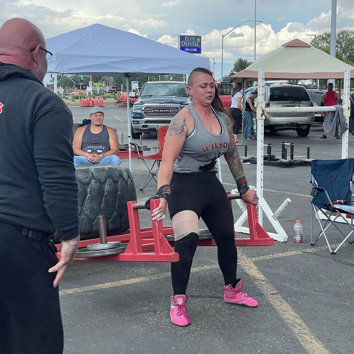 Woman lifting weights in a competition, with a spotter. Outdoors, parking lot setting.